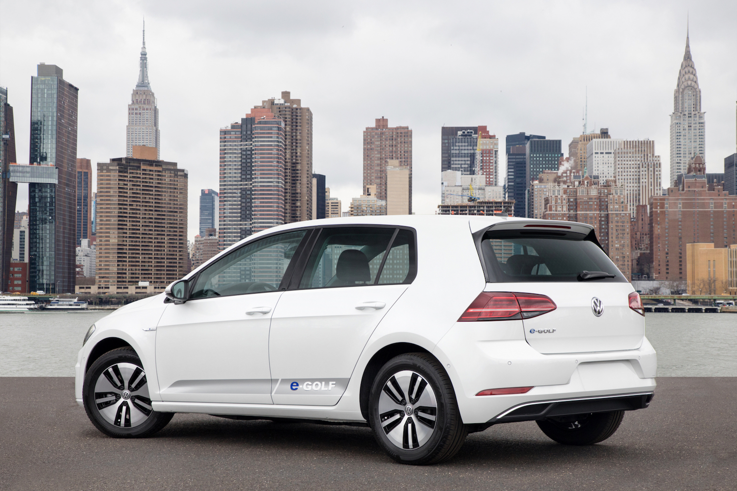 Rear end angle of 2017 Volkswagen e-Golf parked in front of a city with tall buildings.