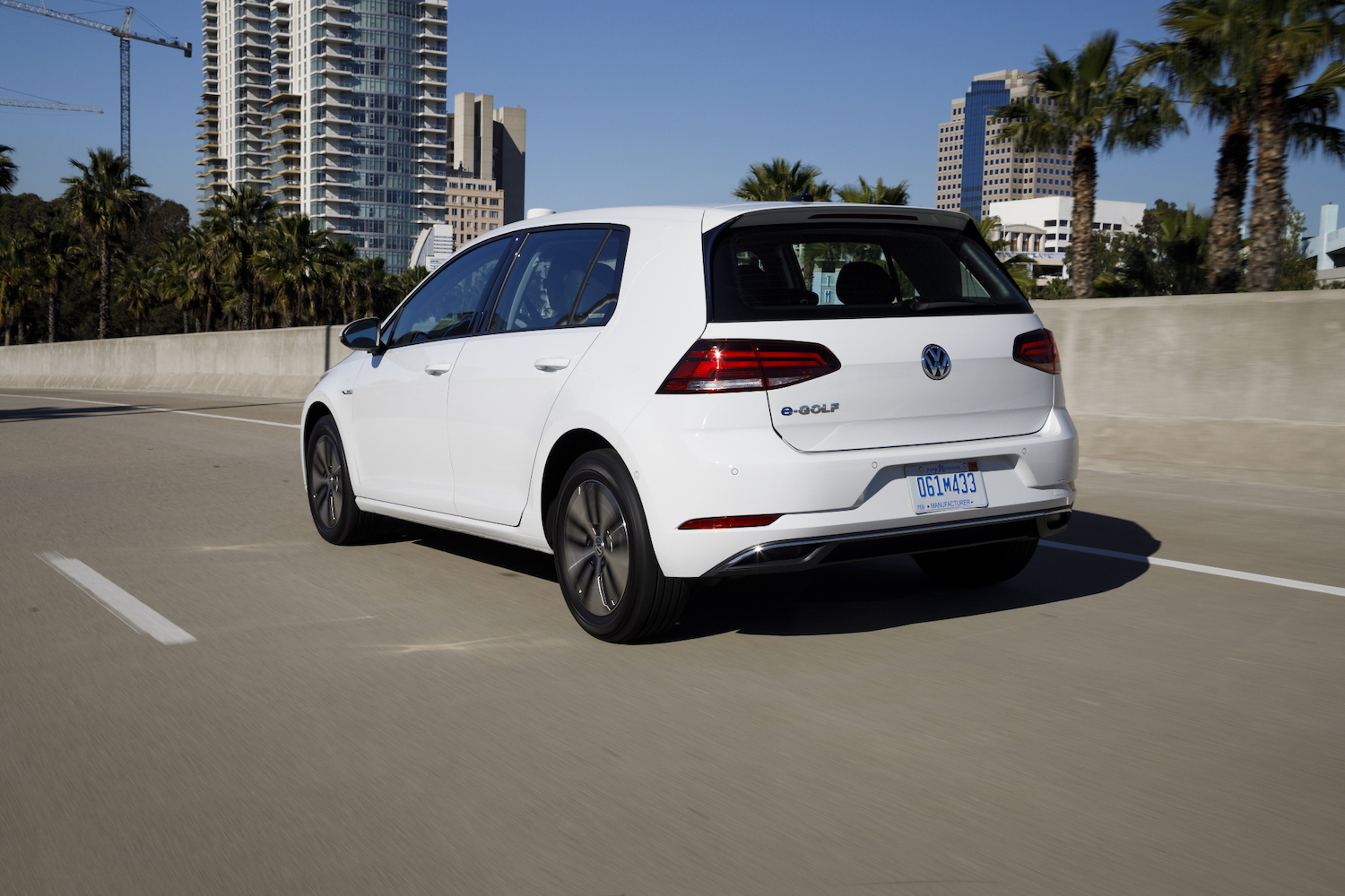 Rear end angle of 2017 Volkswagen e-Golf driving down the road in front of tall buildings.