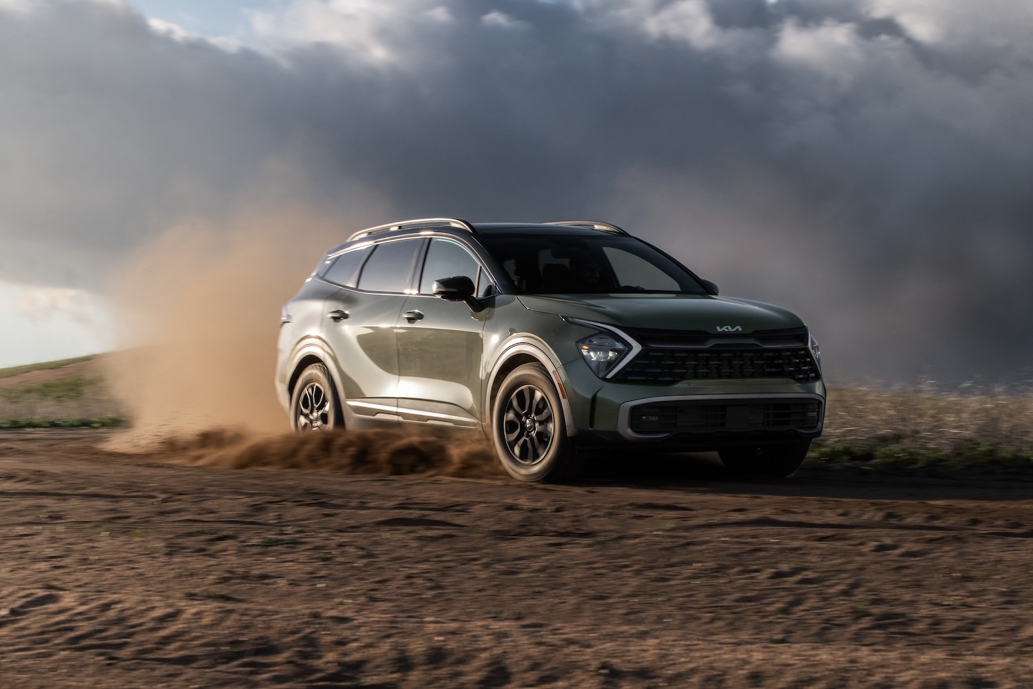 2023 Sportage X-Pro front end angle from passenger side driving on a dirt road with dirt flying up in the back and dark clouds in the sky.