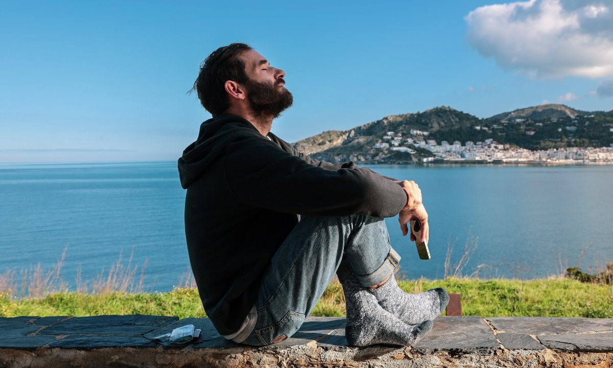 Sad man sitting on the ground near a body of water with his eyes closed.
