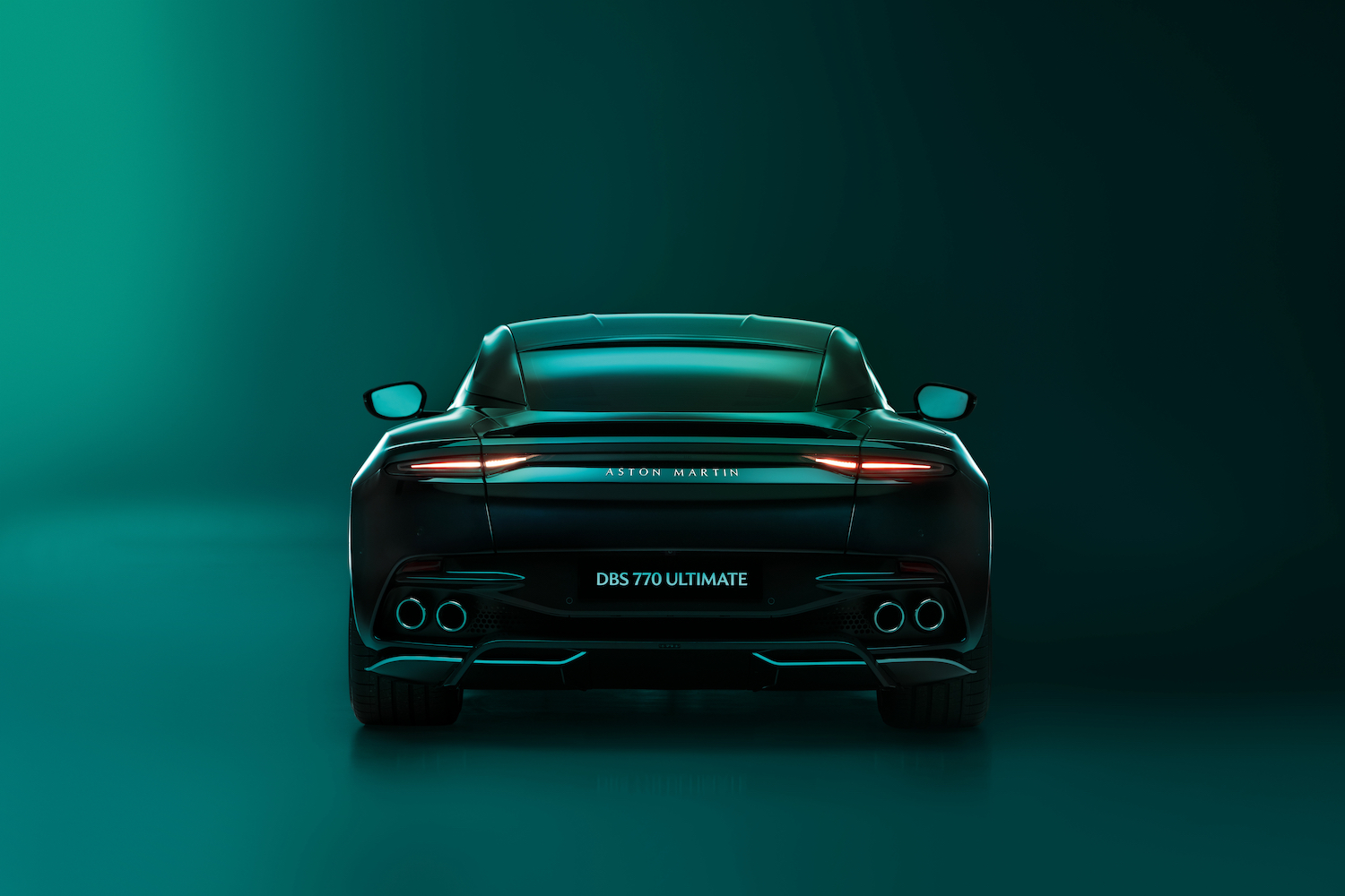 Rear end close up of 2023 Aston Martin DBS 770 Ultimate with taillights on in front of a dark green studio wall.