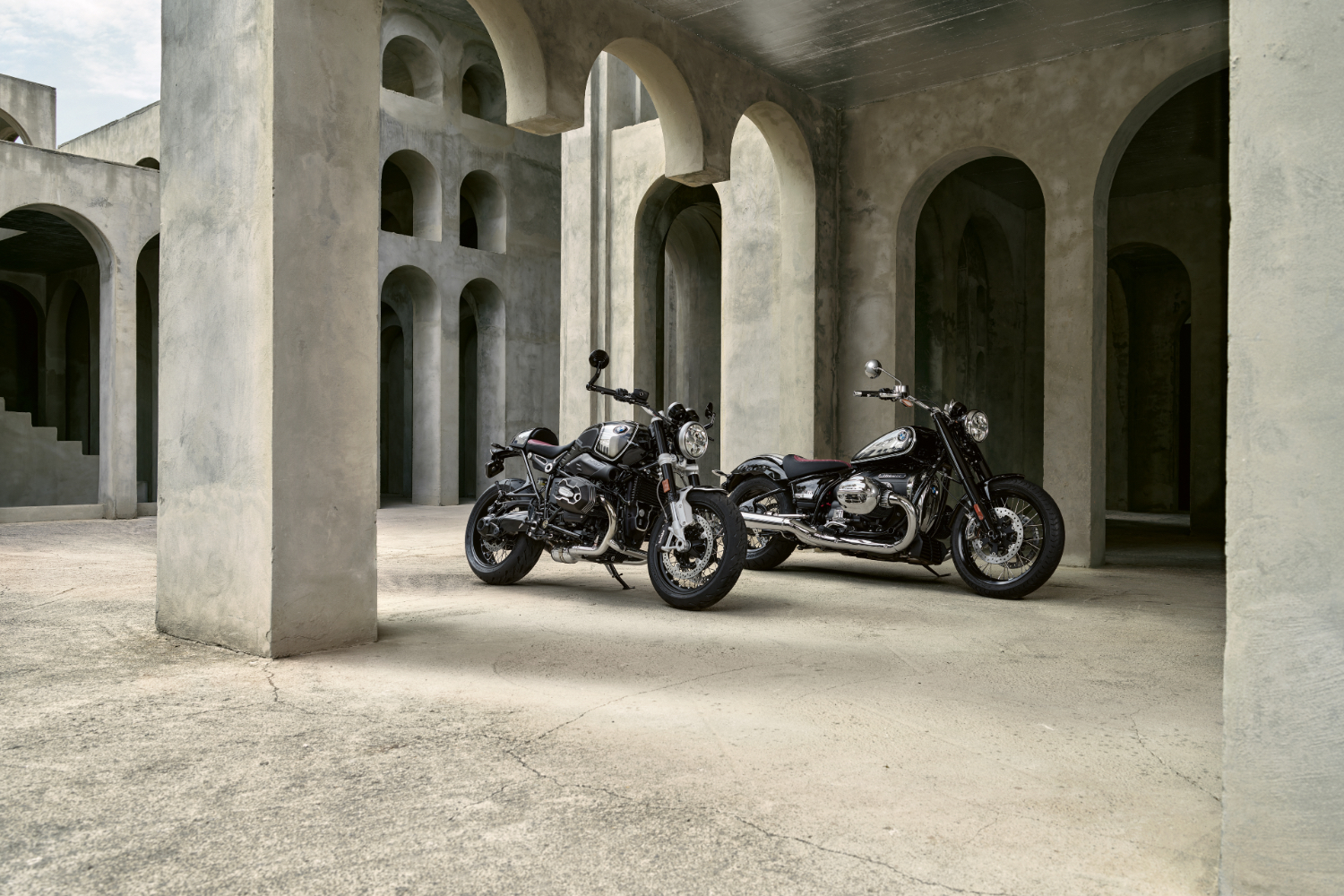 Side angle of 2023 BMW R 18 and R nineT 100 Years Editions parked inside of a villa on top of a dirt path.