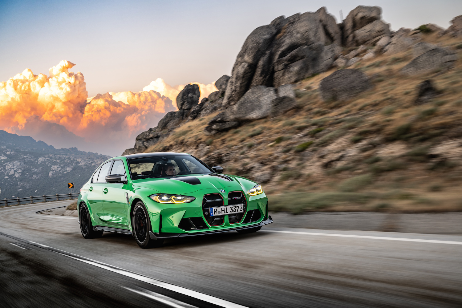 Front end angle of 2024 BMW M3 CS from passenger side driving down a road with mountains in the back.