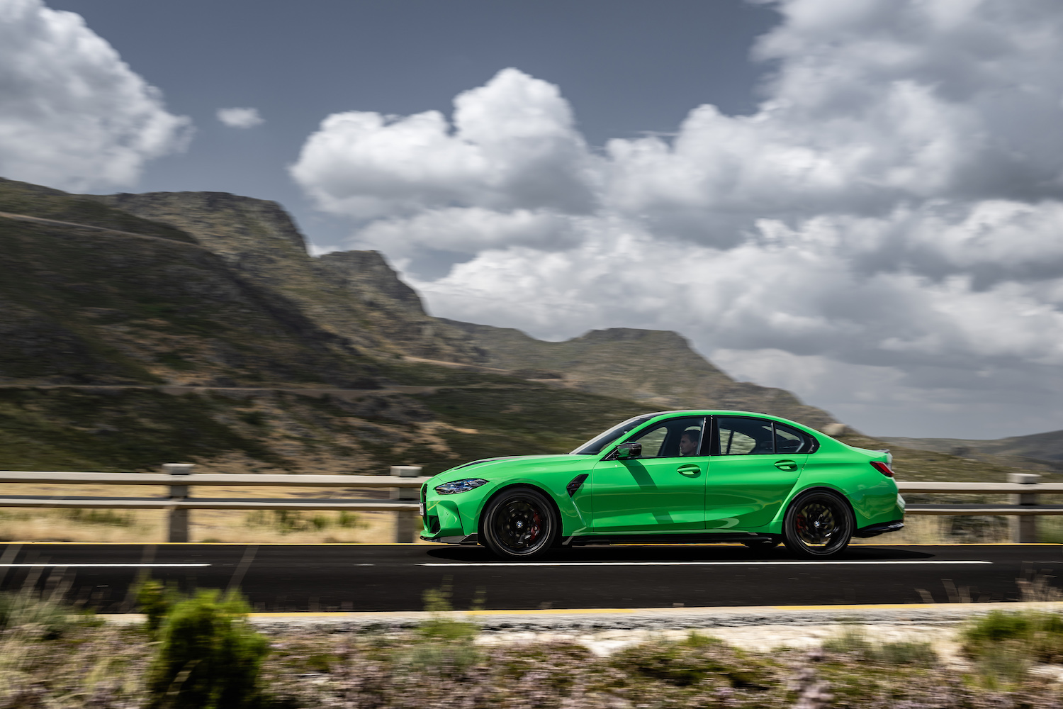 Side profile of 2024 BMW M3 CS driving down a road with mountains in the clouds in the back.