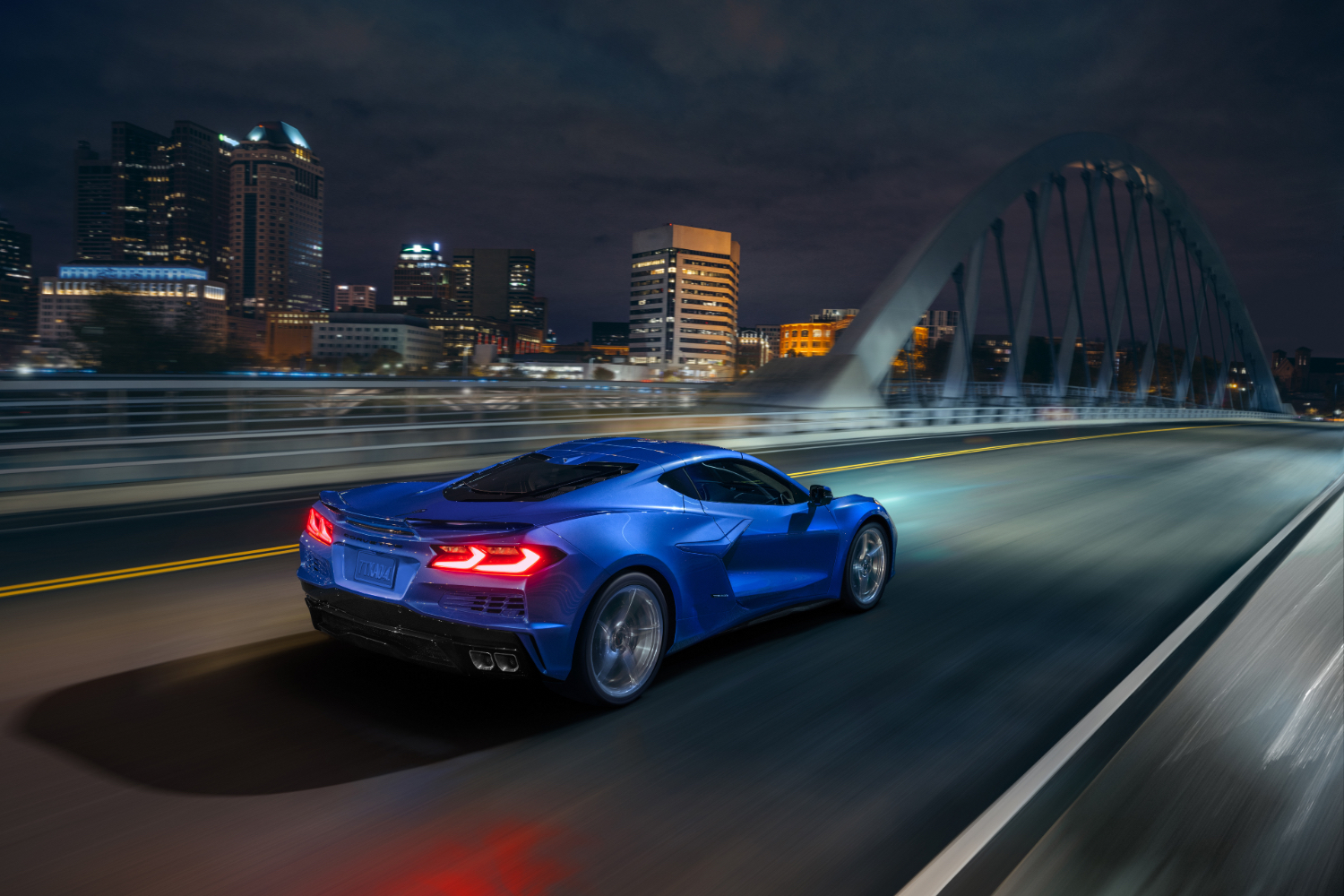 Rear end angle of 2024 Chevrolet Corvette E-Ray driving down the road on a bridge with Detroit city skyline in the back.