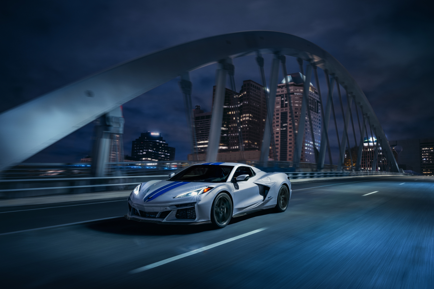 Front end angle of 2024 Chevrolet Corvette E-Ray driving at night down the road with Detroit city skyline in the back.