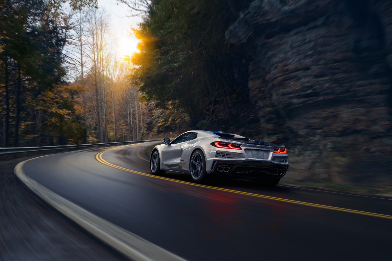 Rear end angle of 2024 Chevrolet Corvette E-Ray driving up a windy road with trees and the sun in the back.