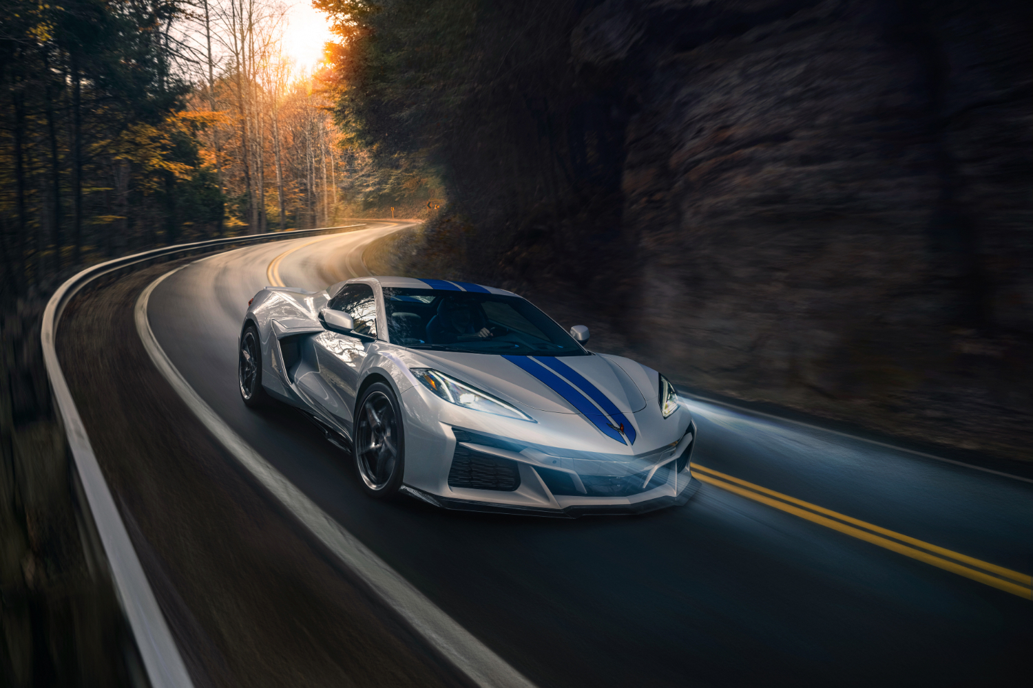 Close up of front end of 2024 Chevrolet Corvette E-Ray driving down the road with headlights on with trees in the back.