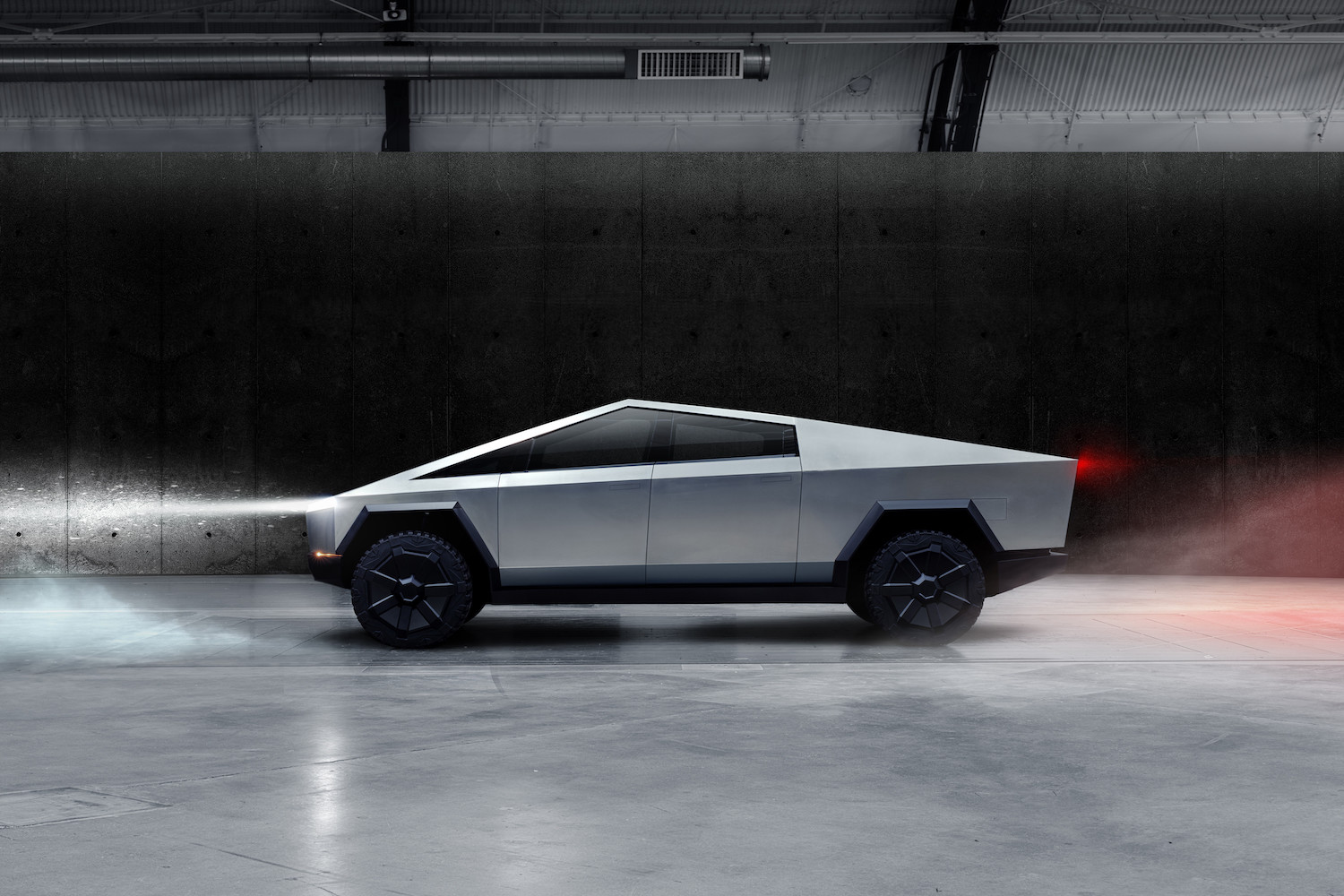 Tesla Cybertruck parked indoors in front of a black wall with headlights and taillights on.