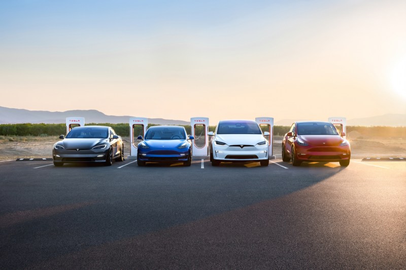 Tesla group photo with Model S, Model 3, Model X, Model Y parked in front of charger during sunset.