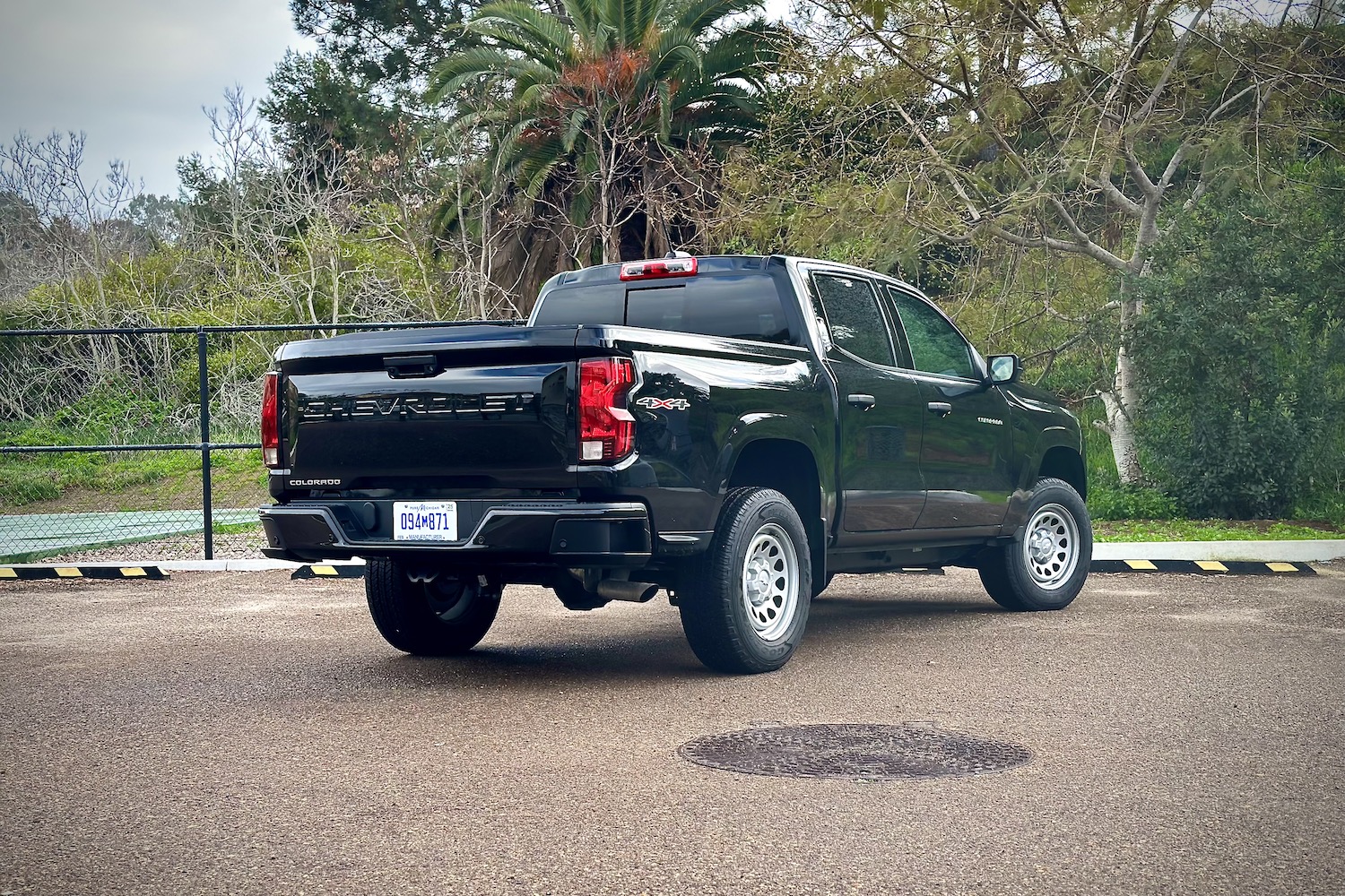 Rear end angle of the 2023 Chevrolet Colorado from the passenger's side parked in front of trees and a tennis court.