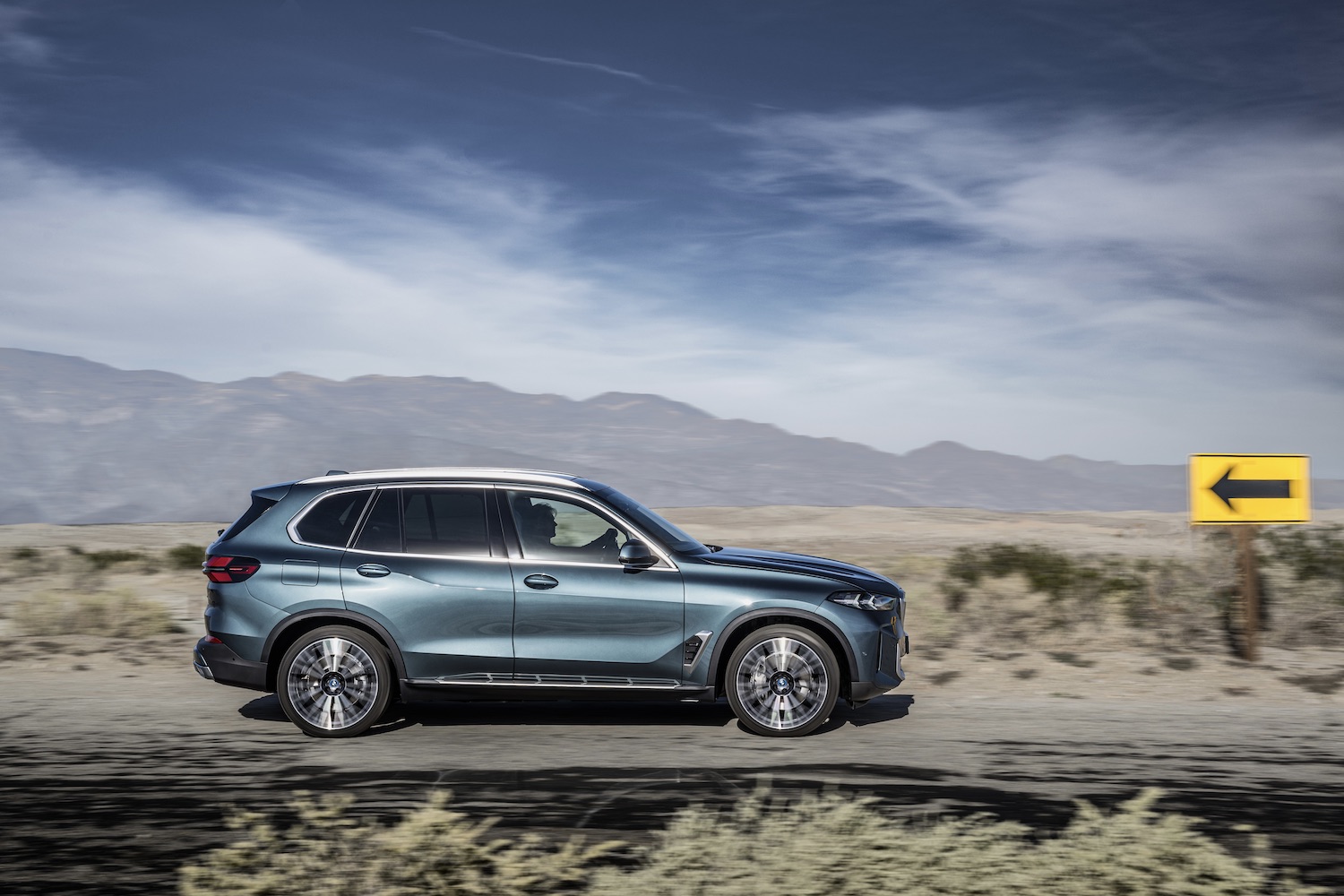 Side profile of the 2024 BMW X5 driving down a road in front of mountains.