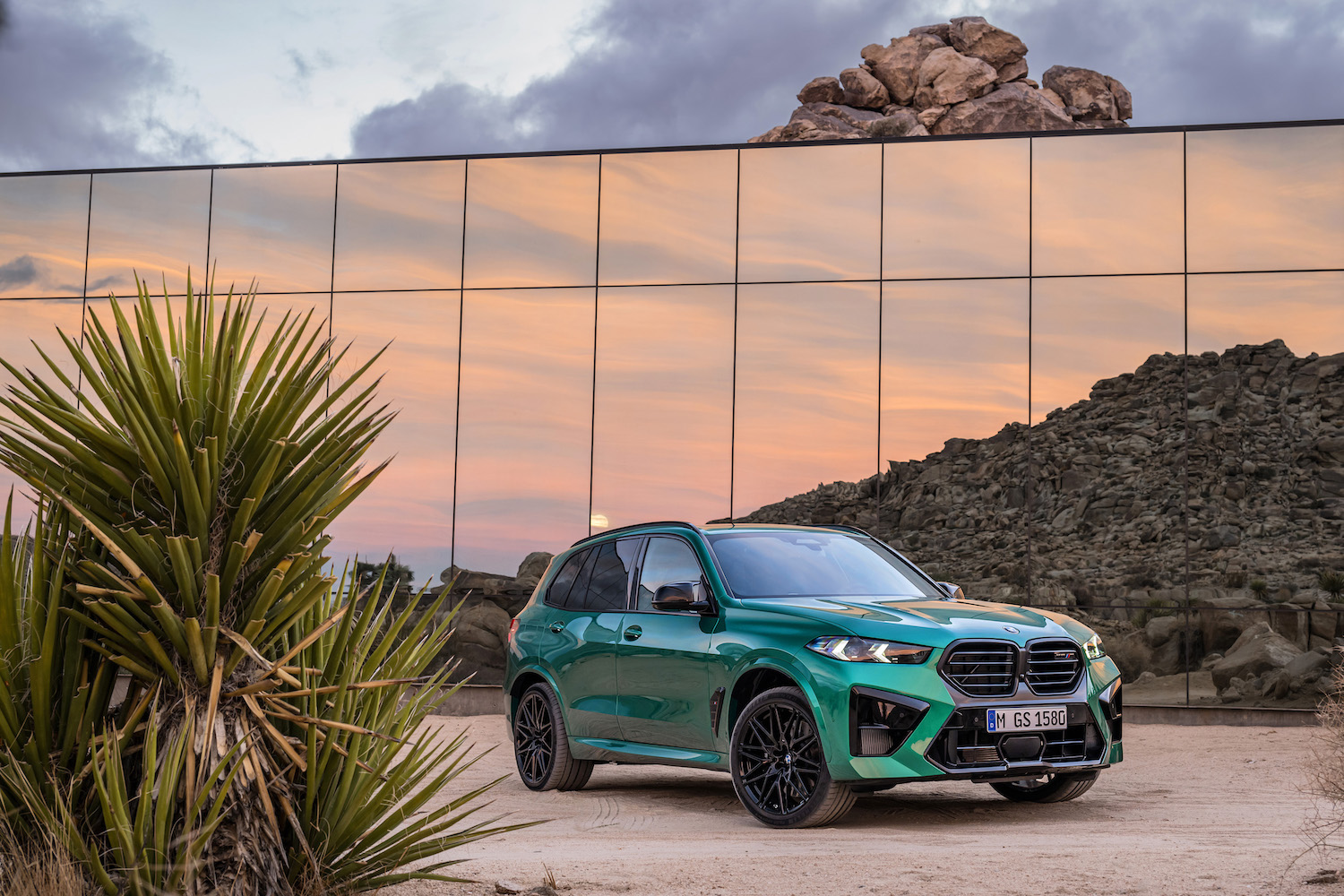 Front end angle of 2024 BMW X5 M Competition from passenger's side parked in front of a glass building.