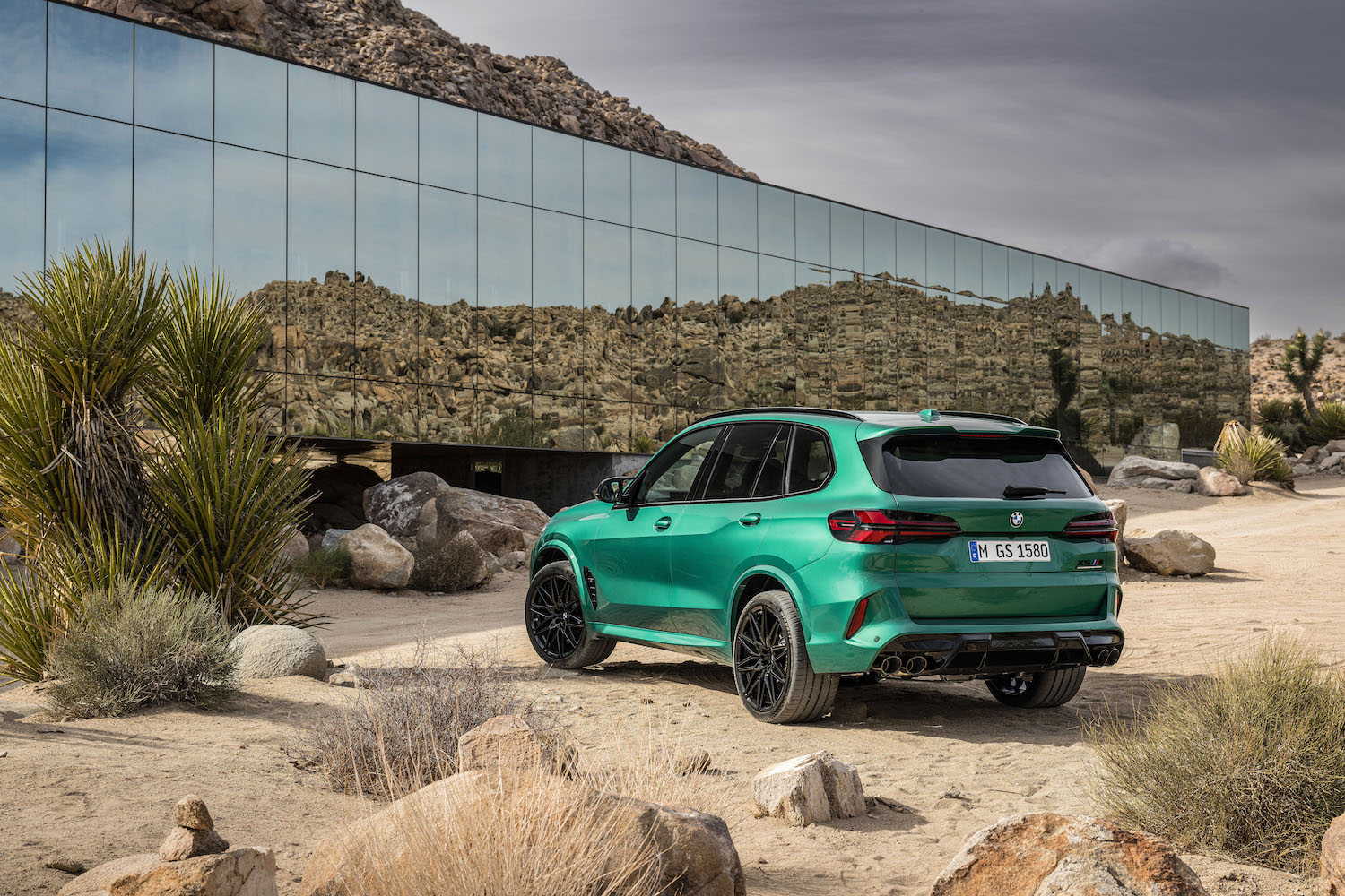 Rear end angle of the 2024 BMW X5 M Competition from the driver's side parked in front of a glass building in the desert.