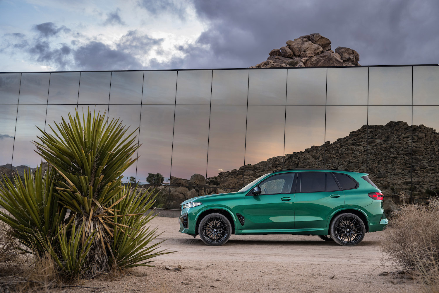 Side profile of the 2024 BMW X5 M Competition parked in the desert in front of a glass building.