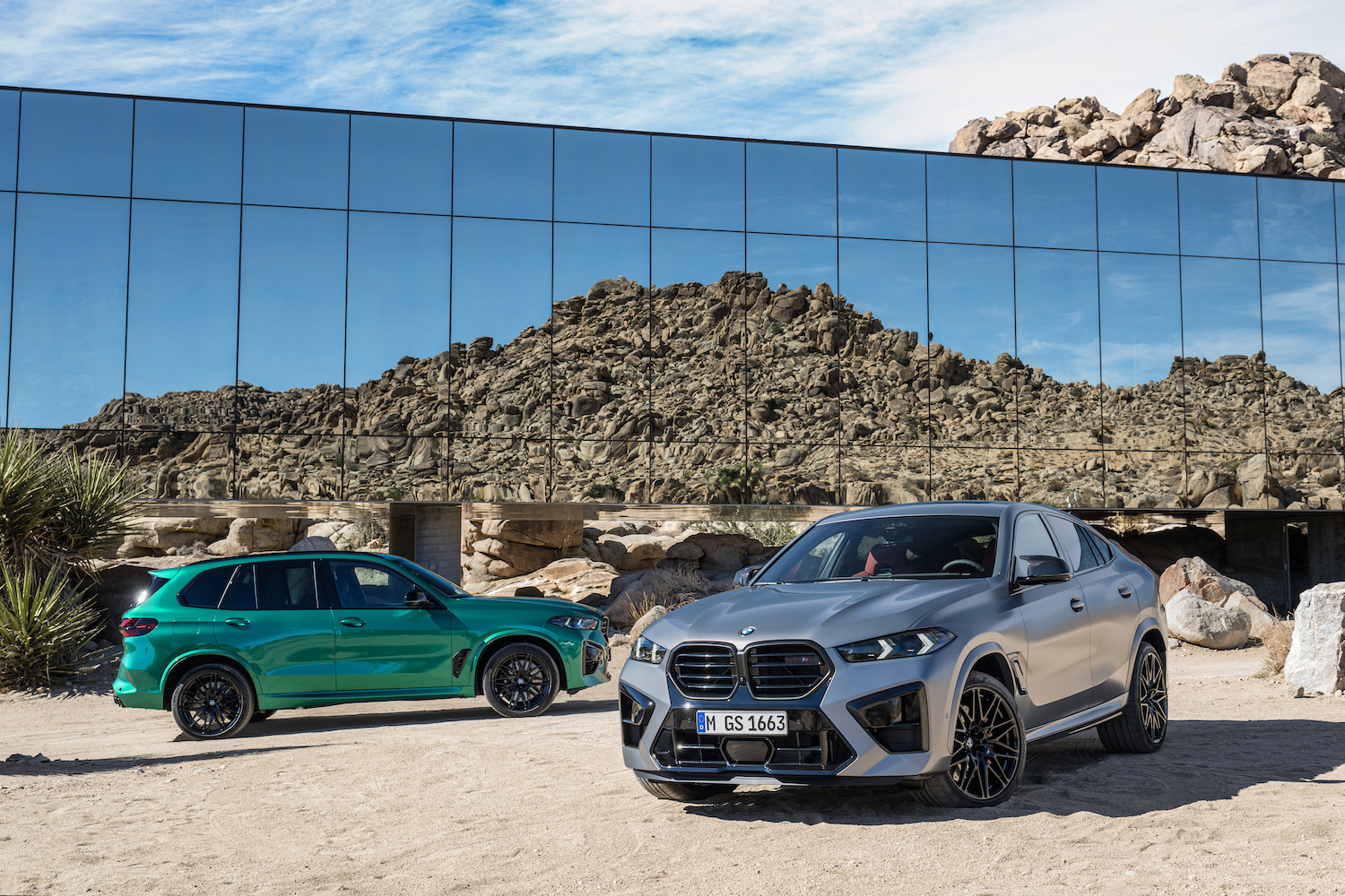 2024 BMW X5 M and X6 M Competition front end angle parked in front of a glass building with mountains in the back.