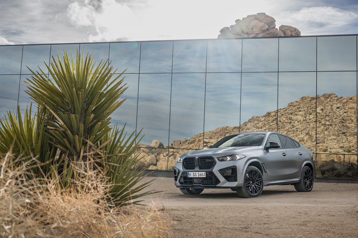 Front end angle of the 2024 BMW X6 M Competition parked in front of a glass building in the desert.