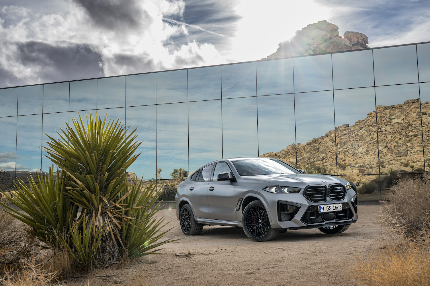 Front end angle from passenger's side 2024 BMW X6 M Competition parked in front of a glass building in the desert.