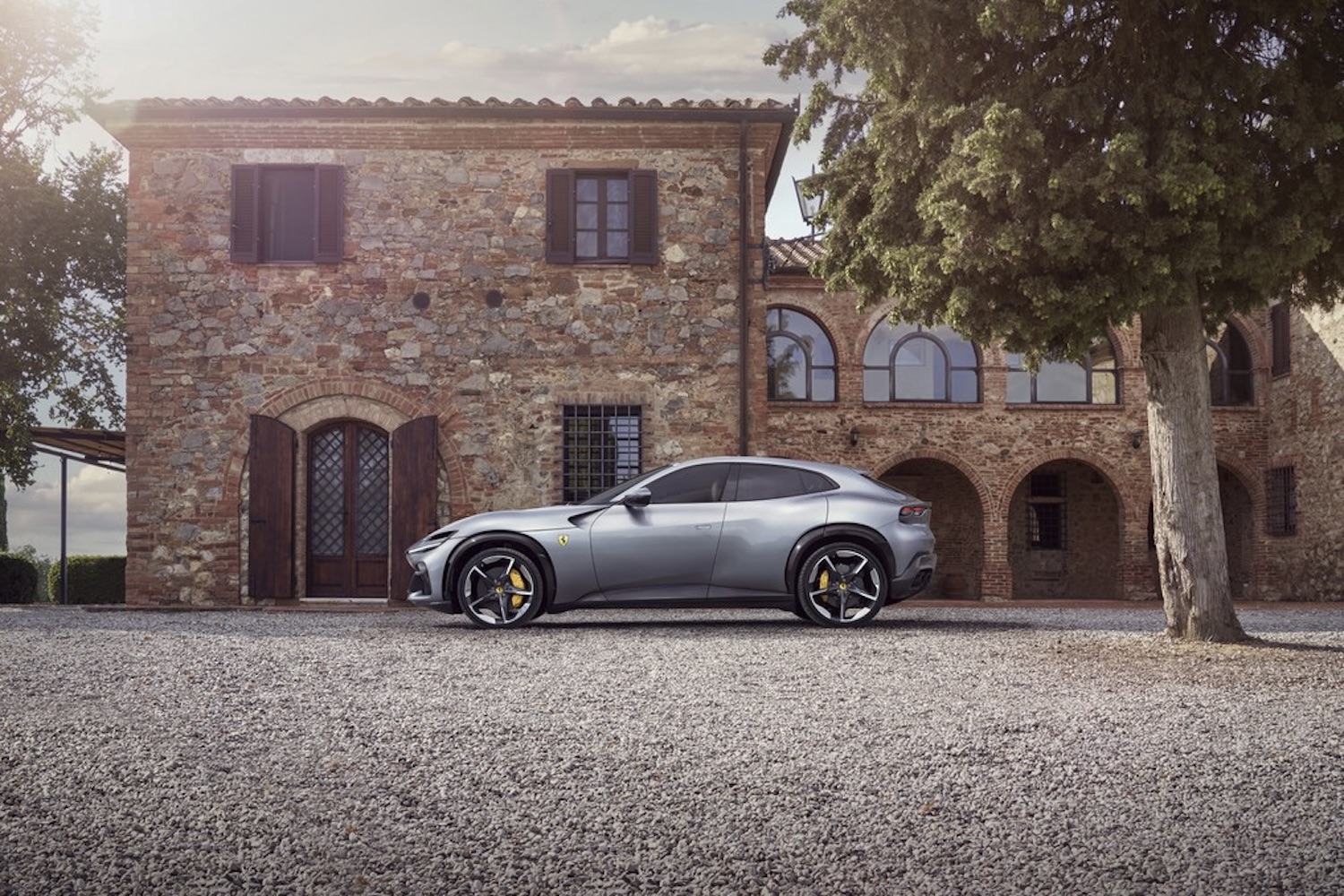 Side profile of the 2024 Ferrari Purosangue parked in front of a stone building with trees in the back.