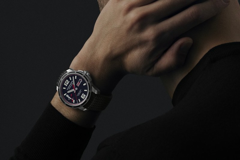 A man wearing a Chopard Mille Miglia GTS Power Control on his wrist in front of a black wall with his hand on his neck.