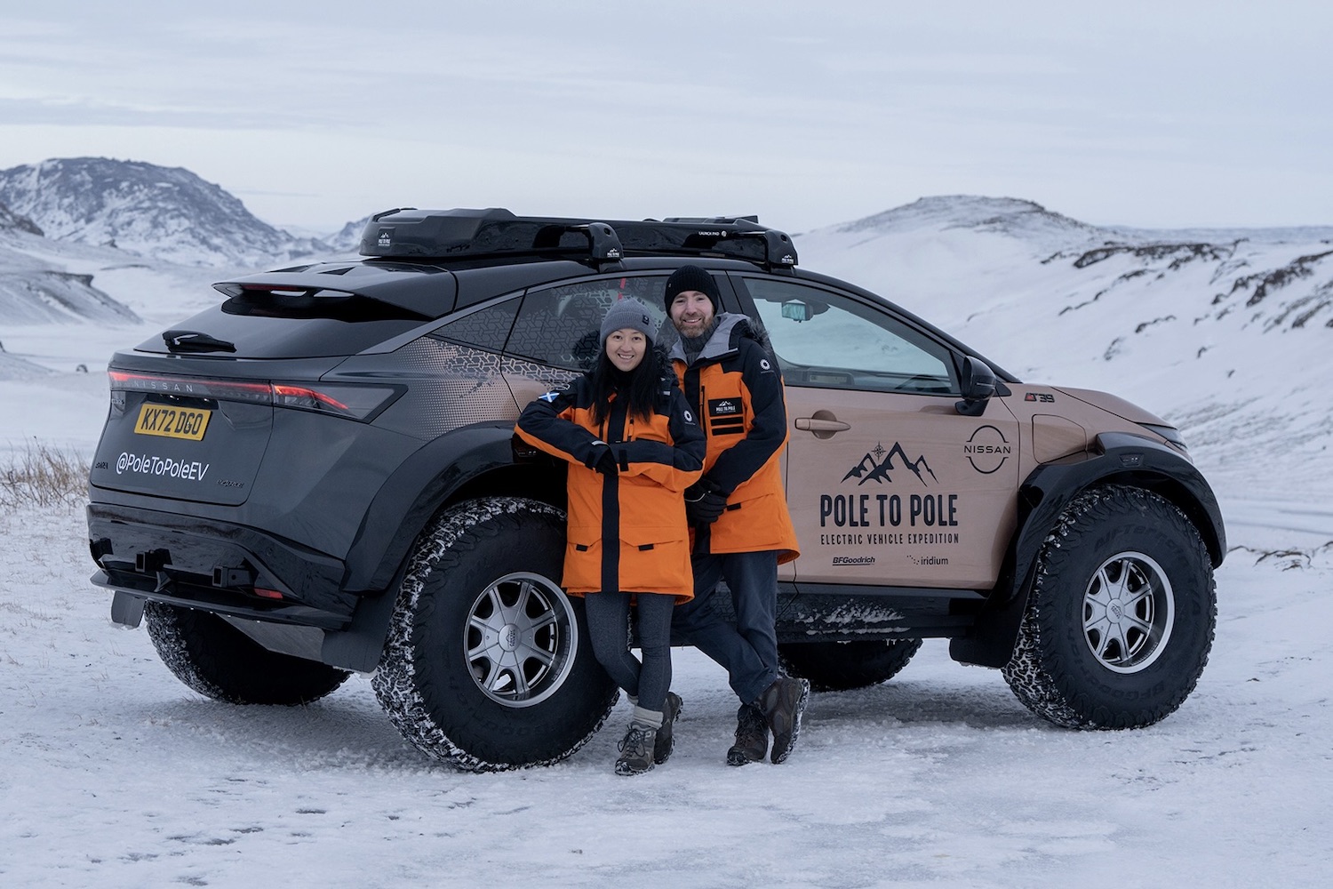 Chris and Julie Ramsey standing in front of Nissan Ariya rear end angle in front of snowy mountains.