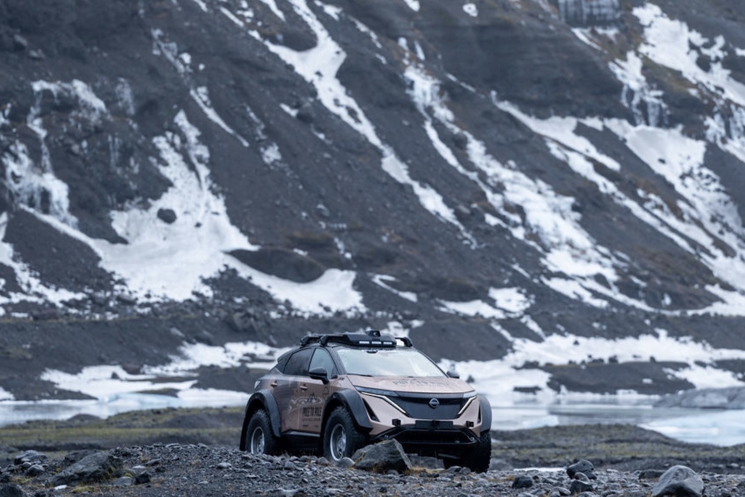 Far away shot of Nissan Ariya parked on rocky terrain with snow and mountains in the back.