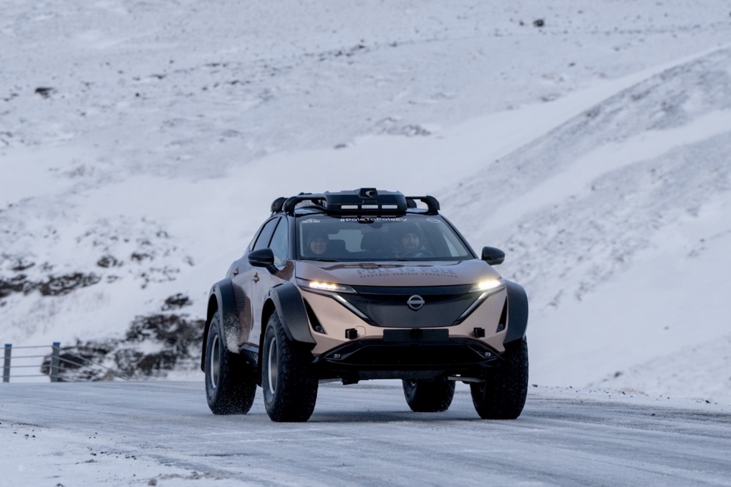 Front end of Nissan Ariya driving down an icy road with snowy mountains in the back.