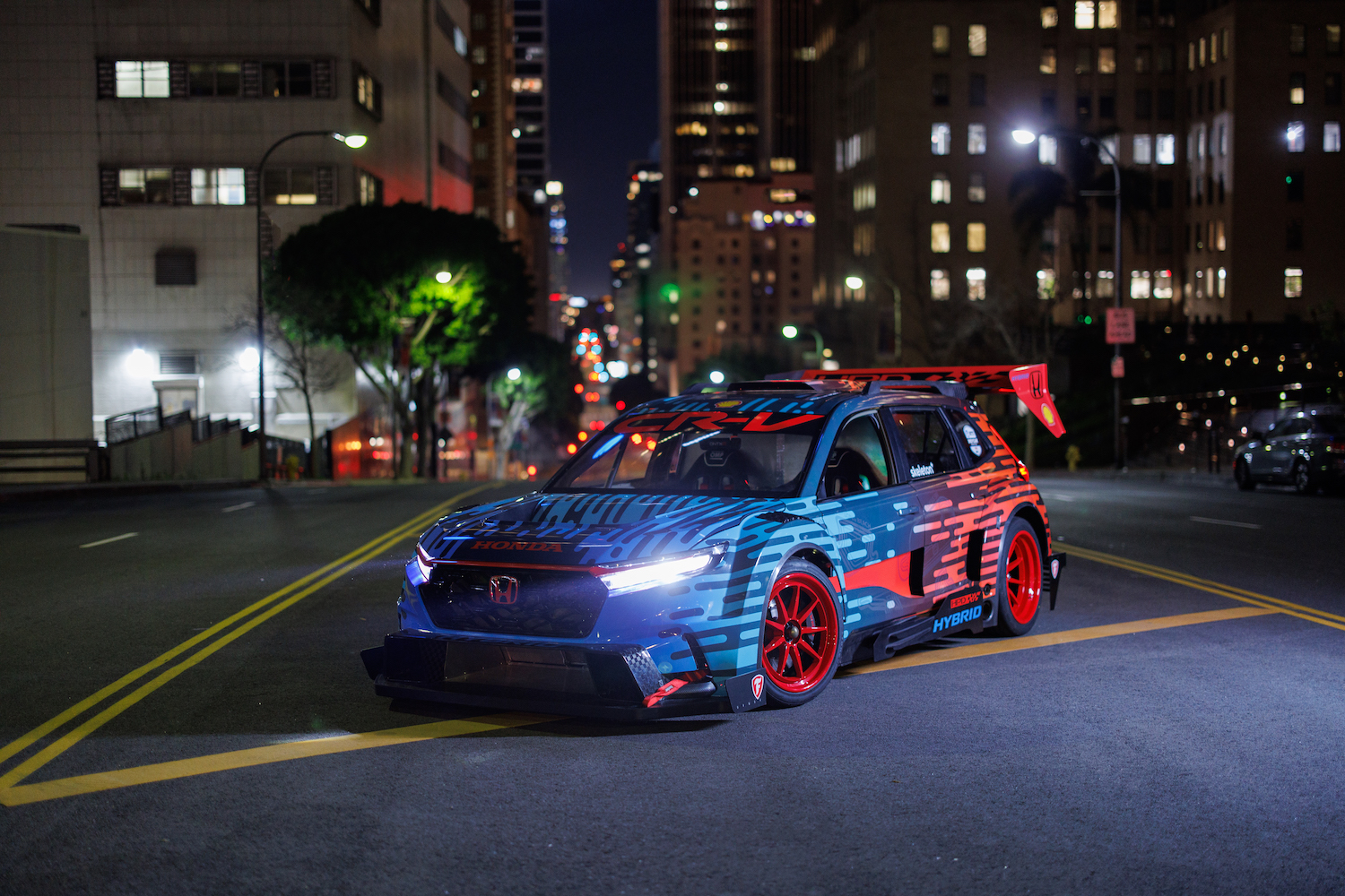 Front end angle of the Honda CR-V Hybrid Racer from driver's side parked in the middle of the road at night in front of tall bui