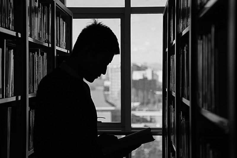 A man in silhouette reads a book between two large bookshelves in front of a window.