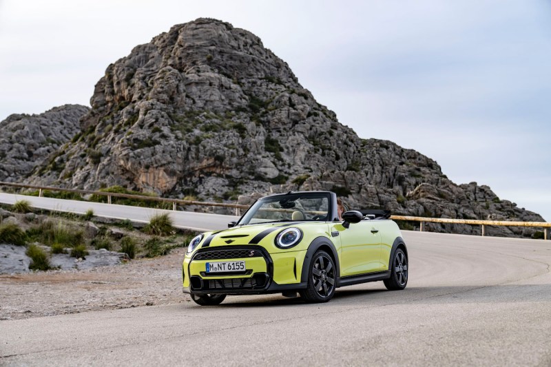 2023 Mini Cooper Convertible parked in front of a winding road and mountain with blue skies.