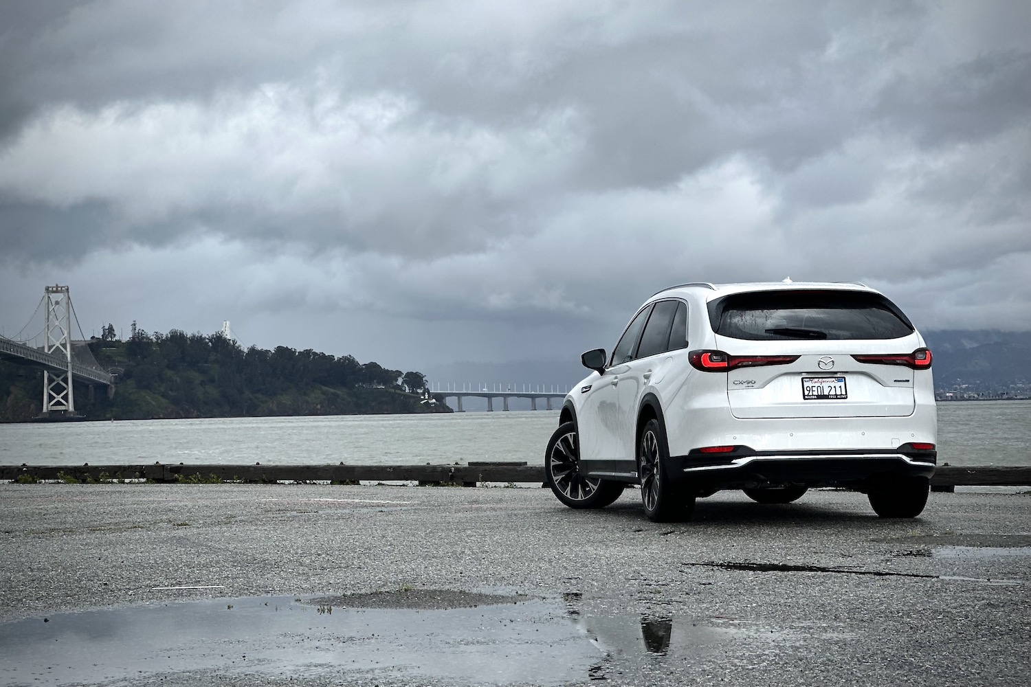 2024 Mazda CX-90 PHEV rear end angle from driver's side in front of water and a bridge.