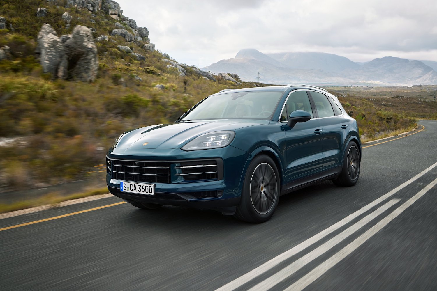 Front end angle of the 2024 Porsche Cayenne driving down the road with a mountain in the back.
