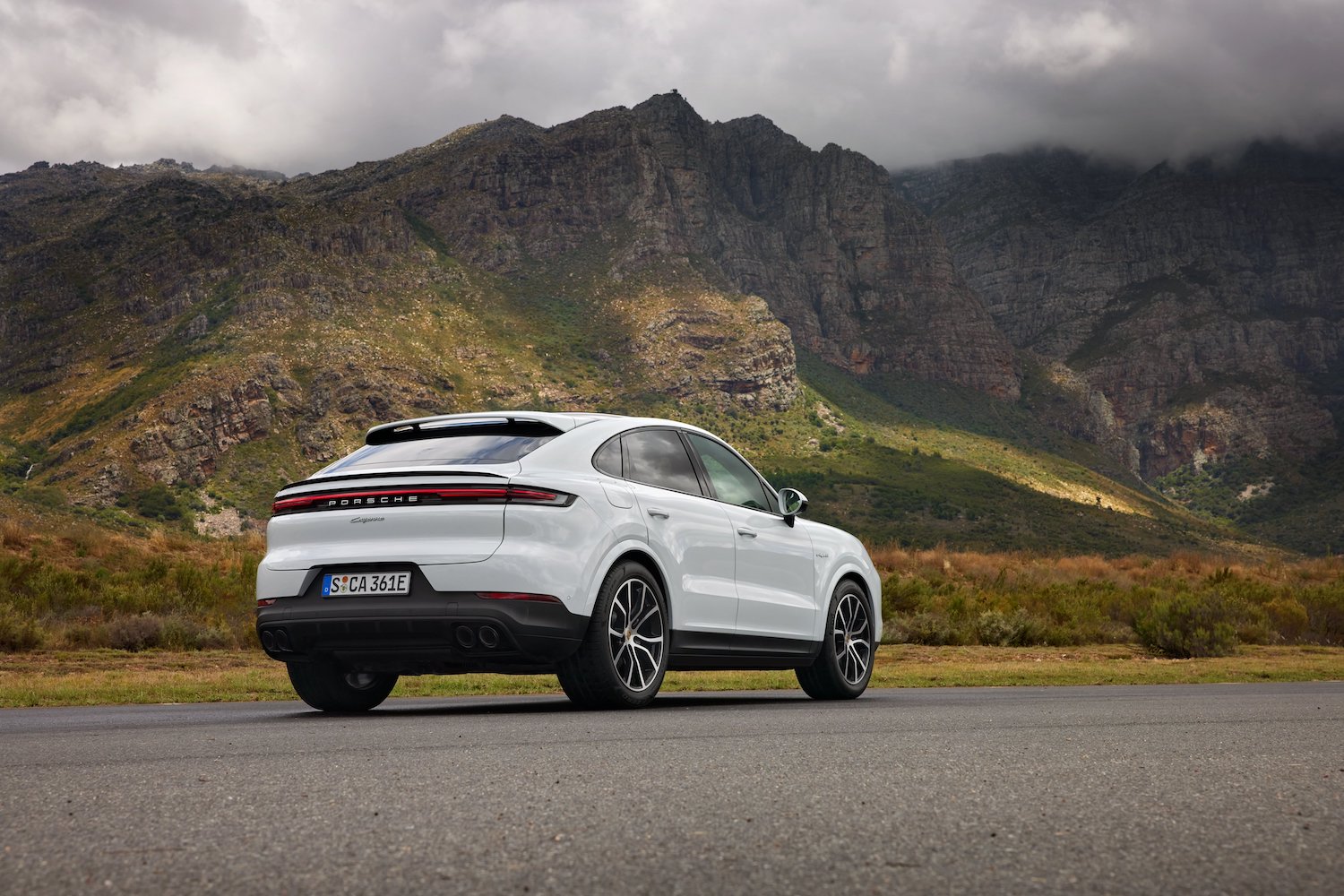 Rear end angle of the 2024 Porsche Cayenne E-Hybrid from the passenger's side in front of mountains.