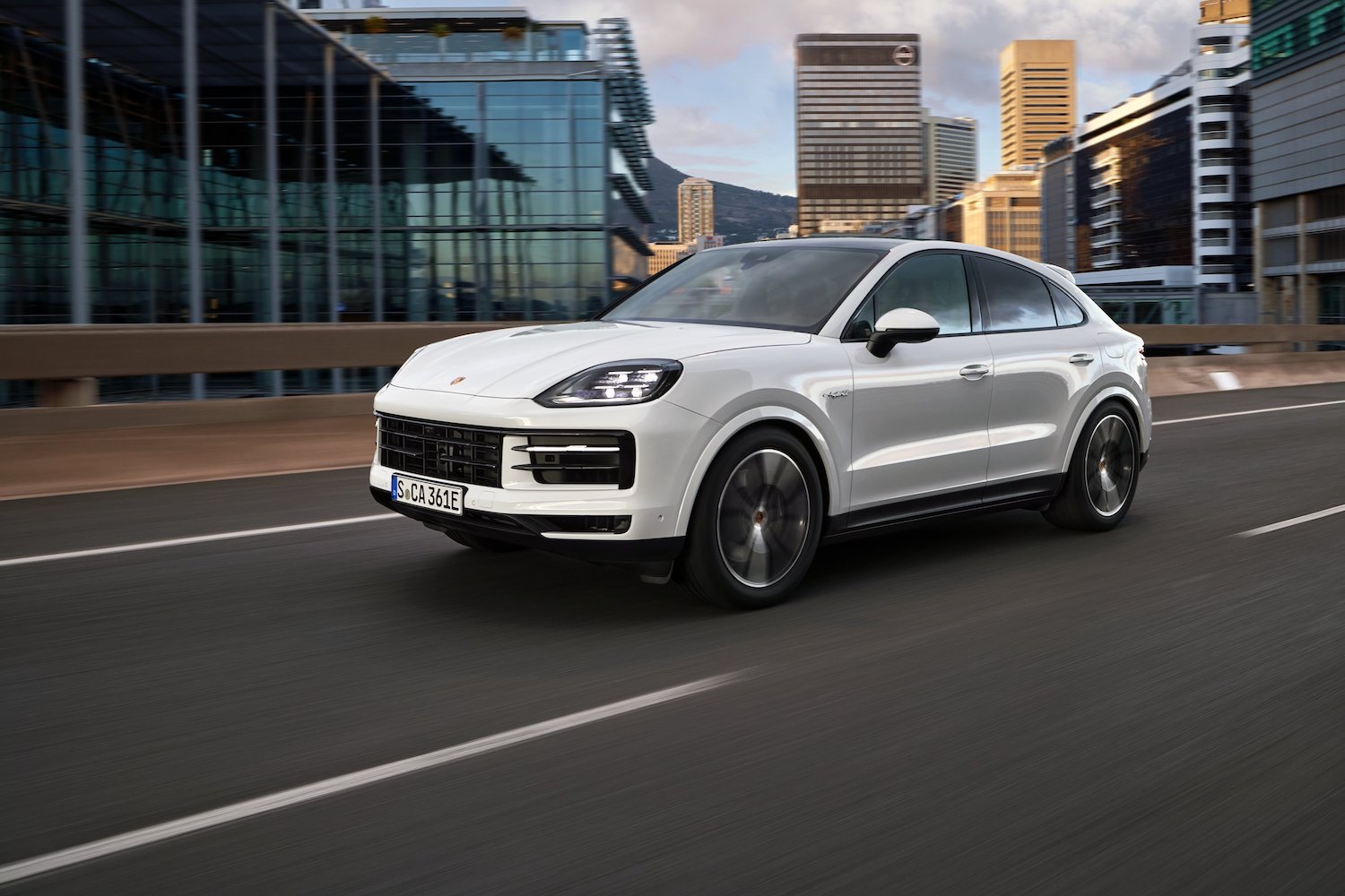 Front end angle of the 2024 Porsche Cayenne E-Hybrid driving down the road in front of buildings.