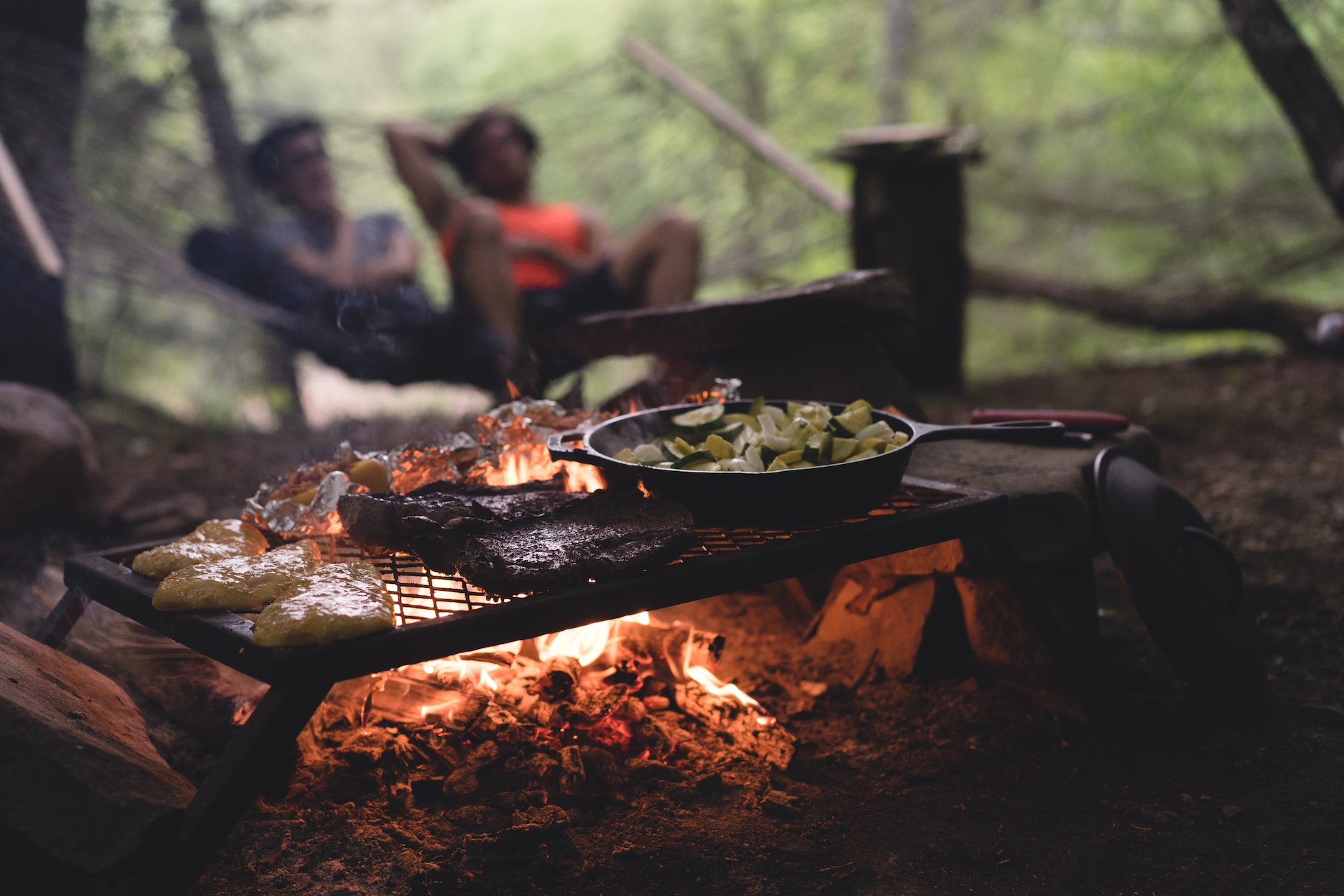 Full dinner grilling on a metal grate over an open campfire with a blurred couple in the background.
