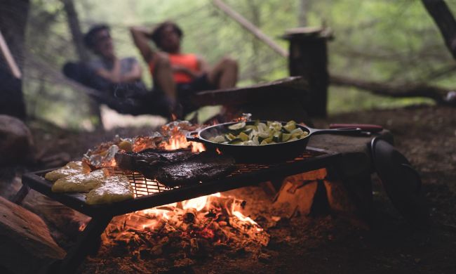 Full dinner grilling on a metal grate over an open campfire with a blurred couple in the background.