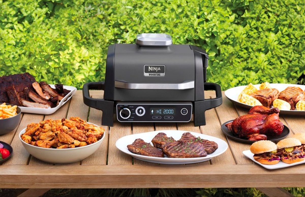 The Ninja Woodfire Outdoor Grill and Smoker on a patio table with plates of food.