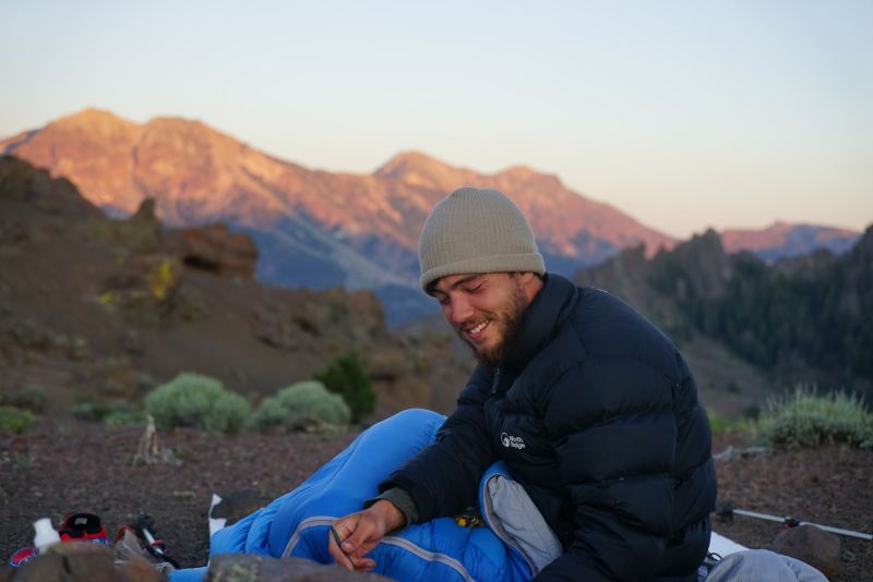 A man sits outside in his sleeping bag overlooking the mountains.