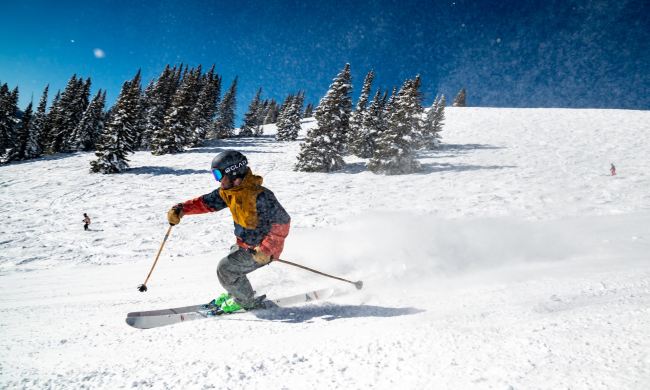 A man skis on a clear run, with mountains and trees behind him.