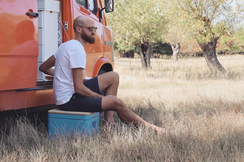 A bearded man sitting on a Trelino portable composting toilet next to an orange campervan.