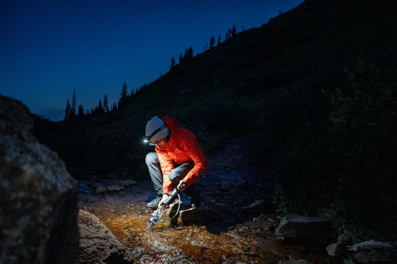 A man collects water from a river by the light of a headlamp.
