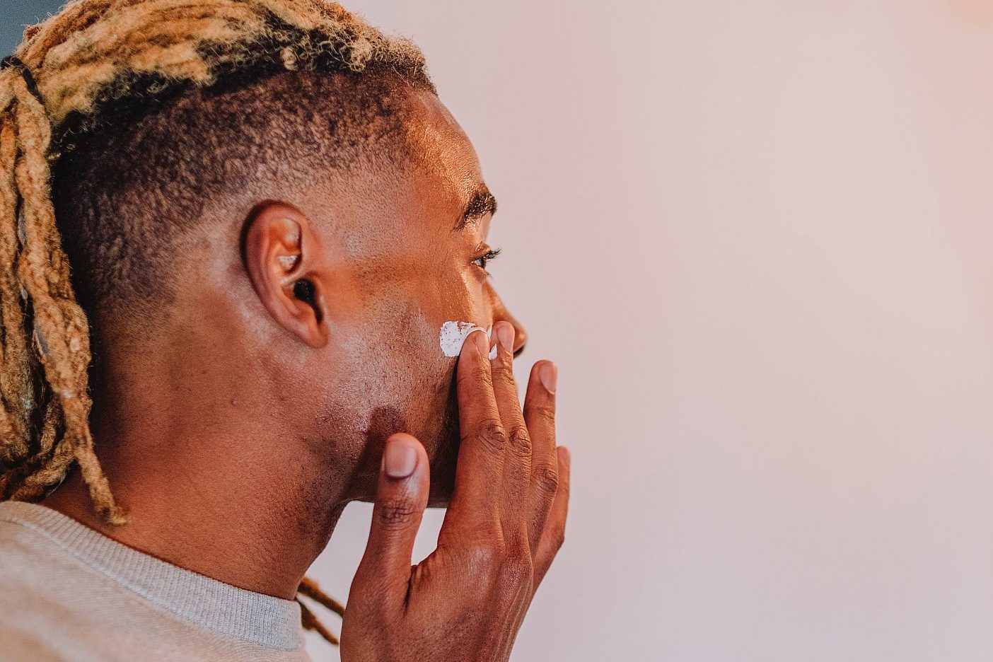 Man with locs applying facial moisturizer in bathroom mirror