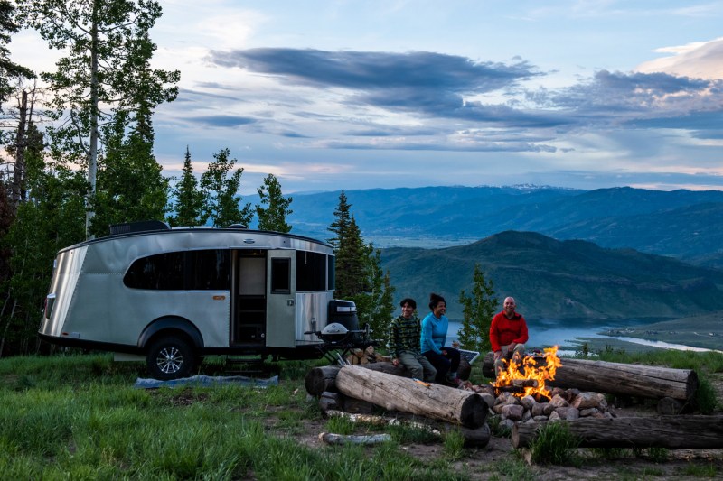 Family of three sitting around a campfire with mountains and an Airstream Basecamp travel trailer in the background.