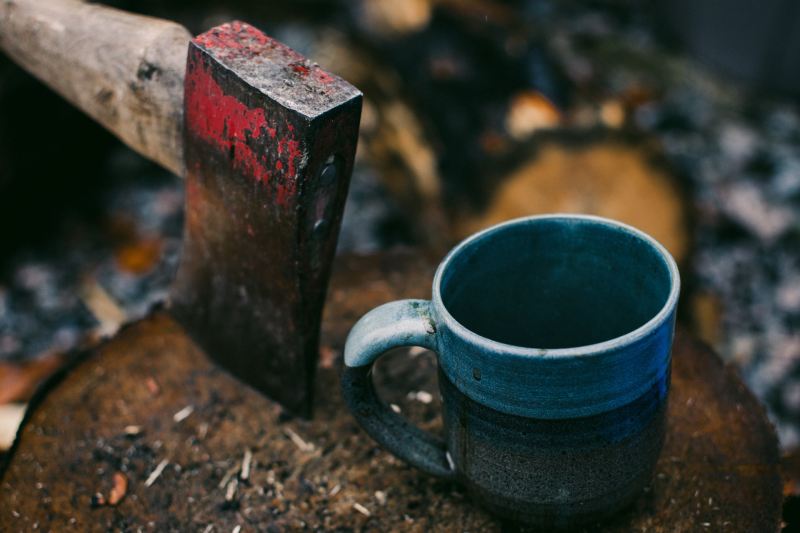 Red-headed ax buried into a tree stump next to a blue coffee mug in the outdoors.