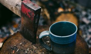 Red-headed ax buried into a tree stump next to a blue coffee mug in the outdoors