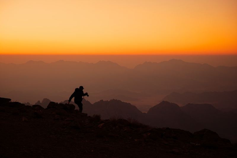 Silhouette of a hiker standing on top of a mountain with a bright orange sky in the background.