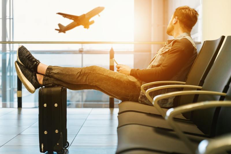 Seated man with feet propped up on luggage looks longingly out airport window at plane taking off