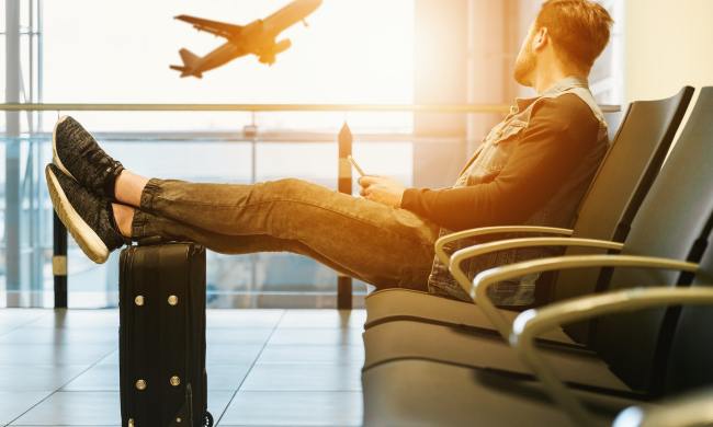 Seated man with feet propped up on luggage looks longingly out airport window at plane taking off