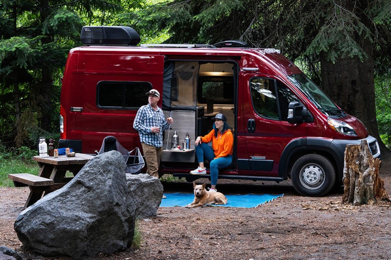 Couple standing outside near the open door of a Winnebago Solis Pocket 36B RV parked at a campsite.