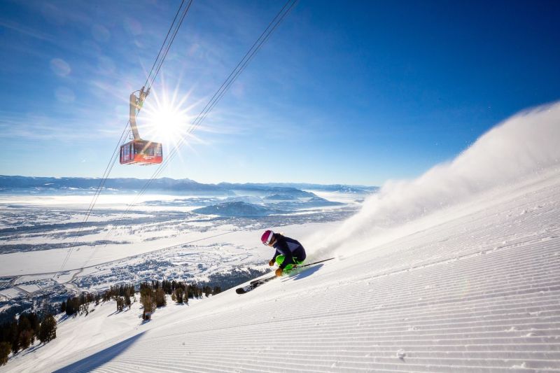 Skier heading downhill at Jackson Hole Mountain Resort, with the sun and aerial tram in the background.